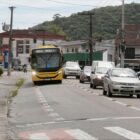 Foto de uma rua com duas pistas de asfalto, um ônibus e um carro em cada uma delas. Há uma calçada com um ponto de ônibus e pessoas embaixo dele.
