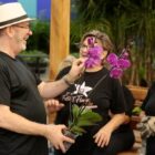 Homem com chapéu segura uma orquídea e sorri enquanto olha para duas mulheres que também sorriem