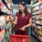 Mulher em corredor de supermercado segura cestinha de compras e um produto