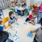 Crianças de uniforme fazem atividade com cartela de letras em sala de aula, em mesas, com professora por perto