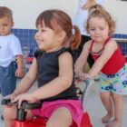 Crianças brincam, uma menina empurra outra numa motoca e outros observam, todas sorriem