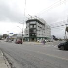 Rua com duas pistas, com carros passando e prédios e casas, onde será implantada a rotatória