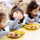 Três crianças uniformizadas estão sentadas em frente a uma mesa e estão fazendo uma refeição com pratos a frente