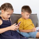 Duas crianças sentadas em sofá mexem em celulares