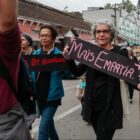 Mulher segura cartaz escrito "mais empatia" e caminha ao lado de grupo de pessoas