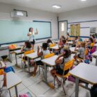 Professora em pé na frente de sala de aula explica para alunos sentados